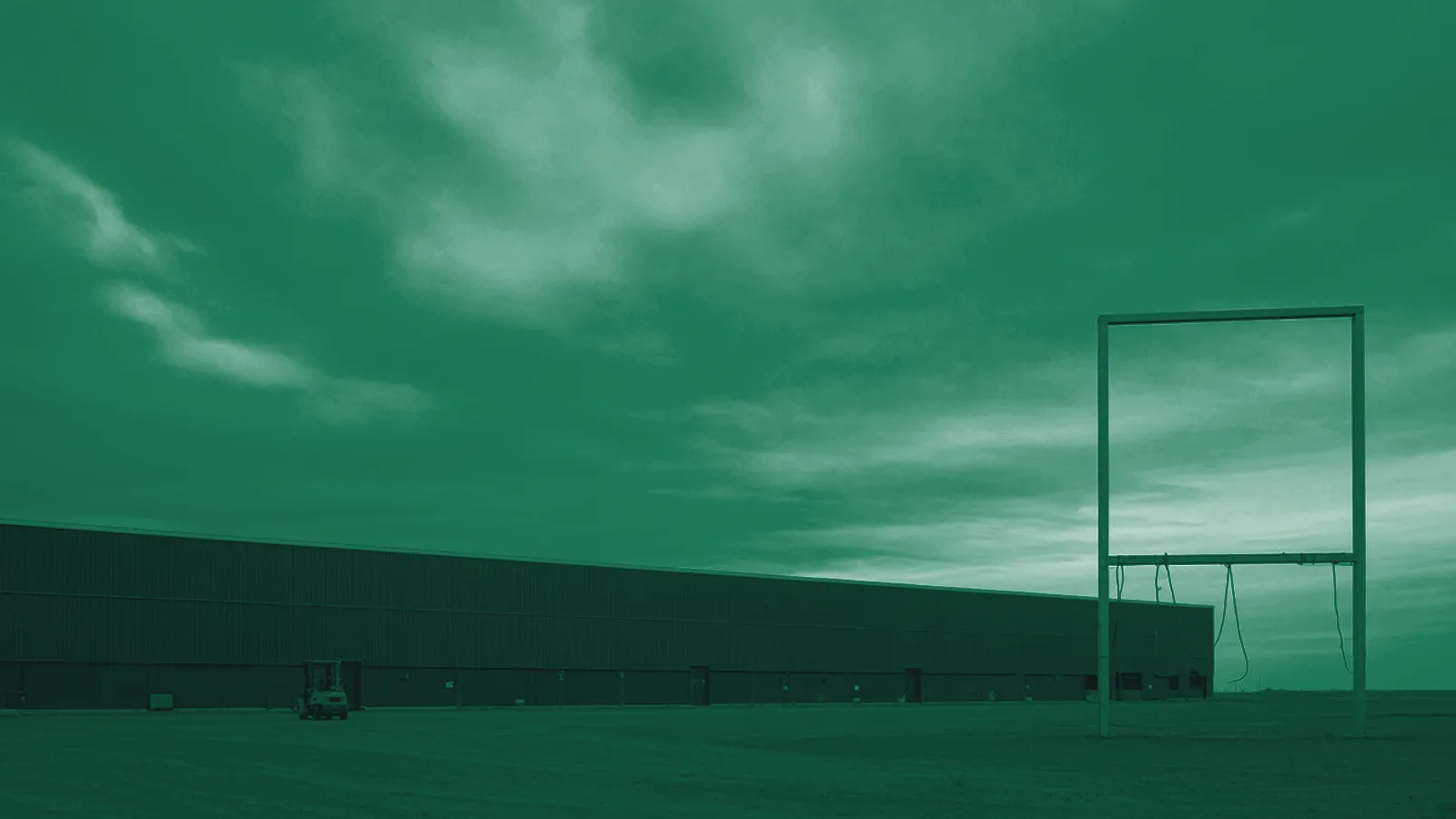 Fotografía de un almacén industrial bajo un cielo nublado, con un panel publicitario vacío sobre postes de acero a la derecha.